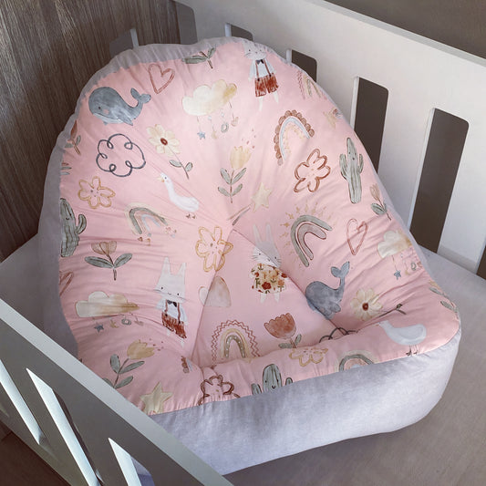 A pink boho-style baby lounger with a pattern of animals and plants, placed in a wooden crib.