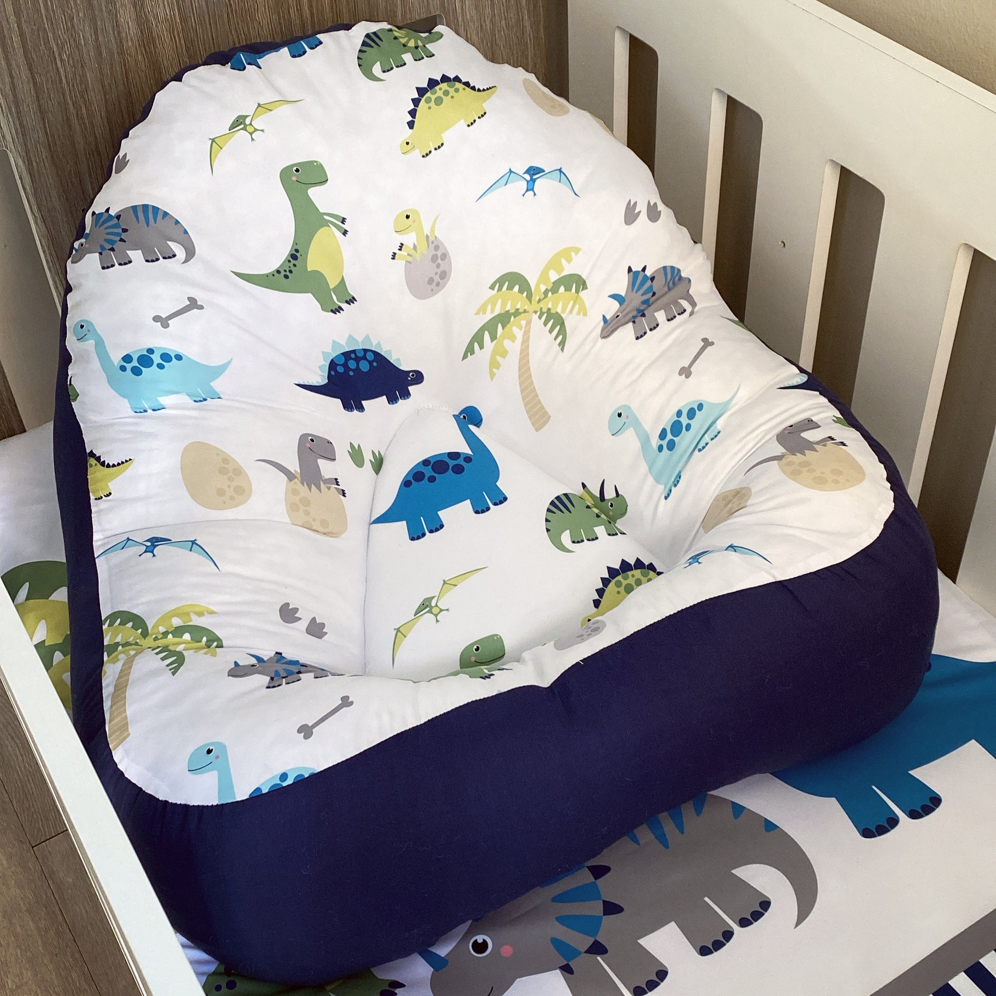 A dinosaur-themed baby lounger with a predominantly white fabric featuring colorful dinosaur prints and a solid blue cushion base.