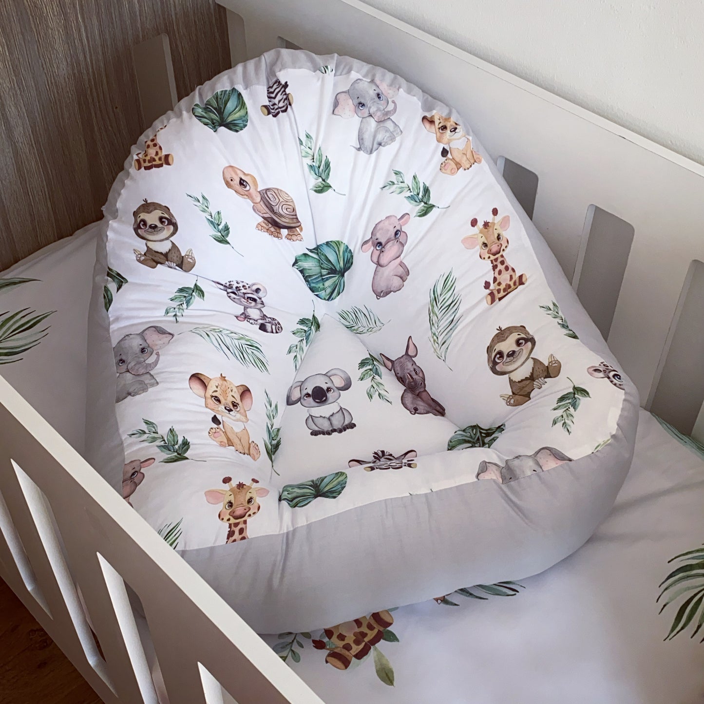 A safari-themed baby lounger with a white fabric featuring colorful animal illustrations, placed in a white crib.