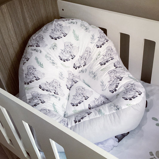 A white fabric lounger with a sheep pattern, positioned in a wooden crib.
