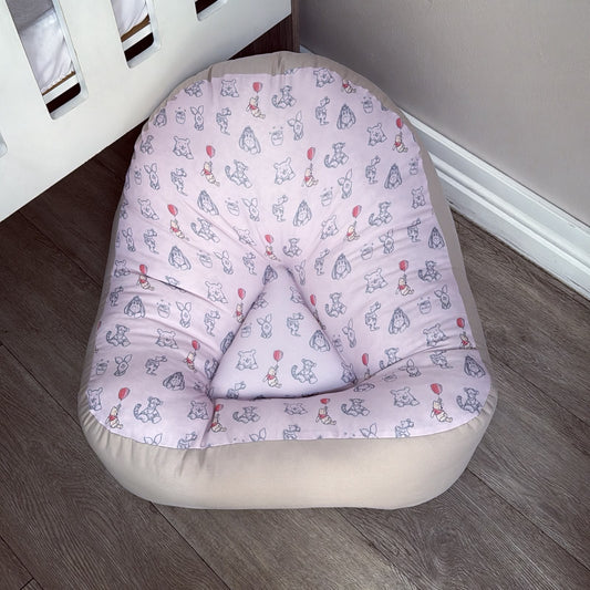 A baby lounge chair with a light pink and white patterned cushion and a beige fabric base, placed on a gray wood floor next to a white crib.