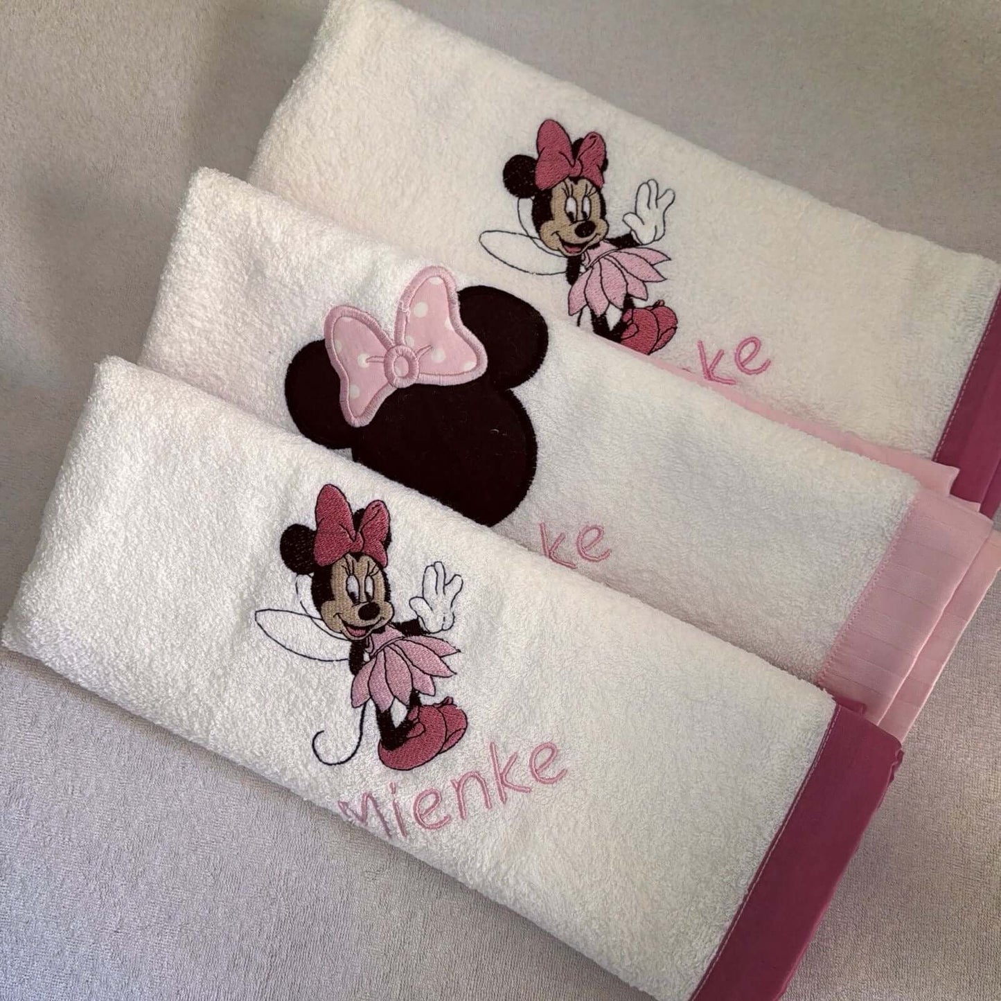 Two white towels with Minnie Mouse designs and personalized names on a gray surface.