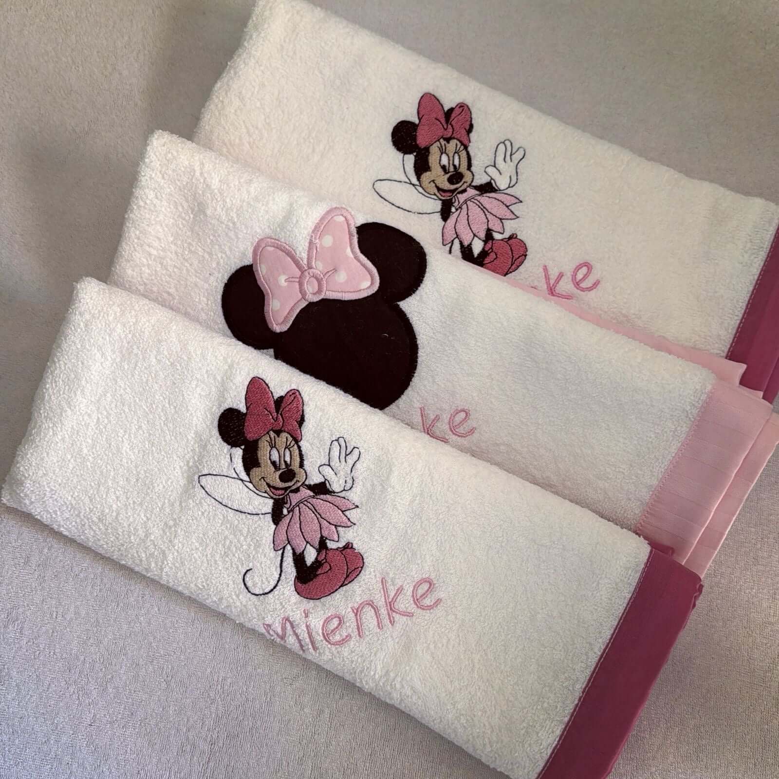 Two white towels with Minnie Mouse designs and personalized names on a gray surface.