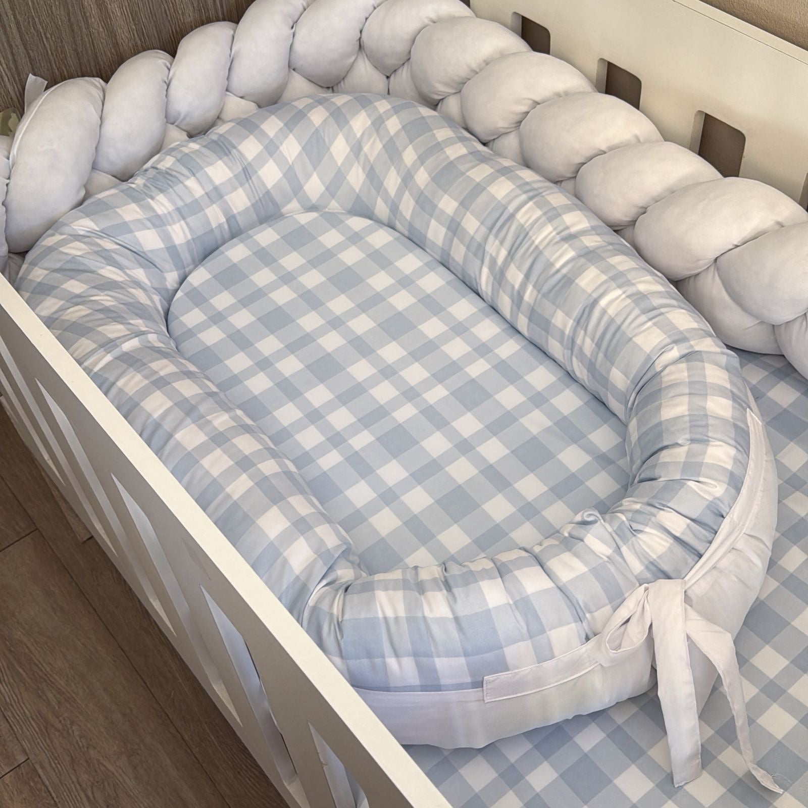 Baby crib with a blue and white checkered bumper pad on a wooden floor.