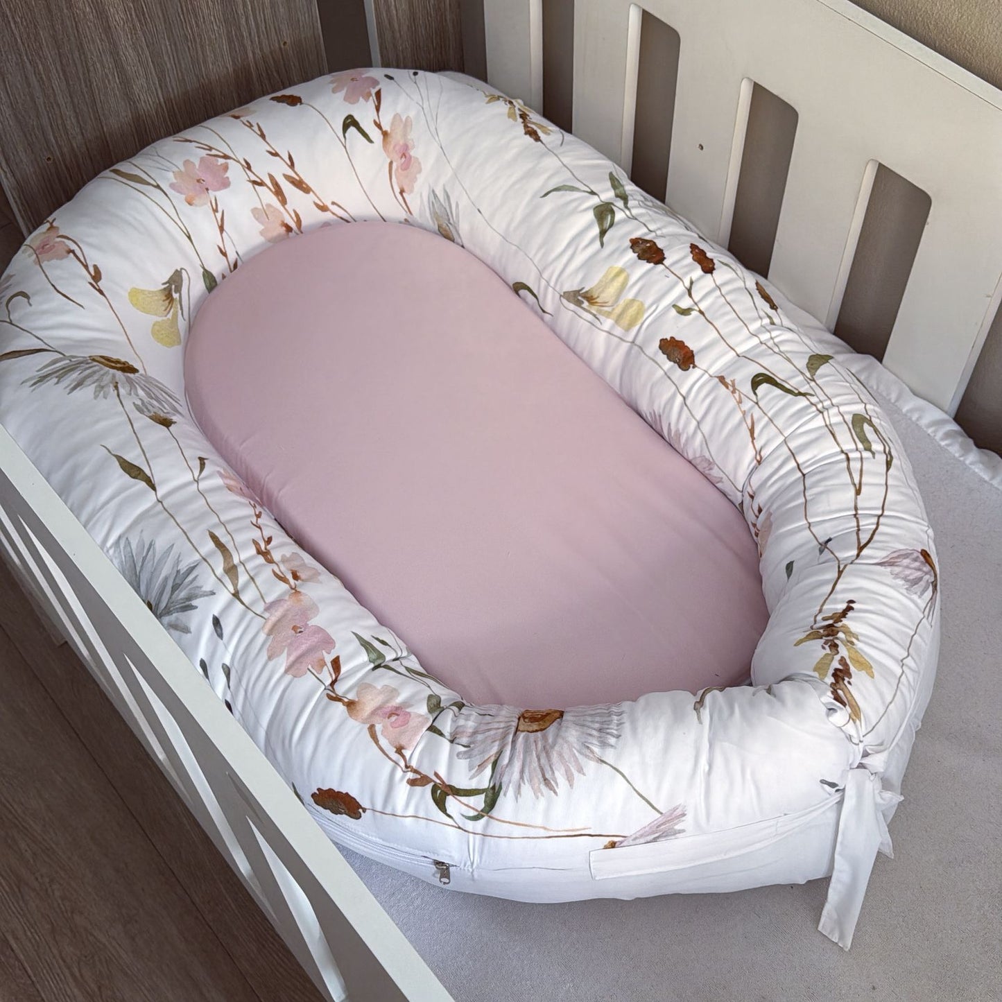 Floral-patterned baby nest with a pink cushion inside a crib.