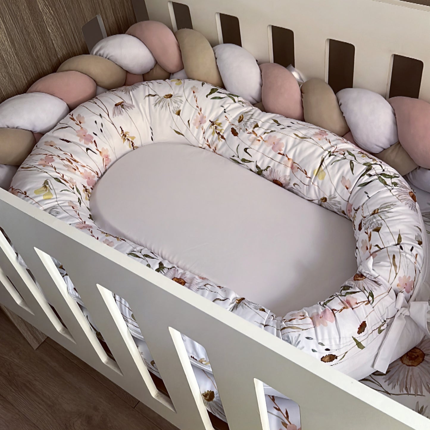 A floral-patterned baby nest placed inside a crib, surrounded by a variety of soft, round cushions.