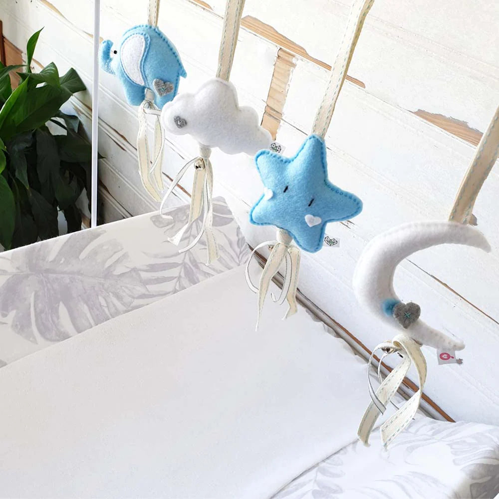 A baby crib hanging toy set featuring a blue star, white cloud, blue whale, and white crescent moon in soft felt fabric, suspended from wooden crib bars over a white leaf-patterned mattress.