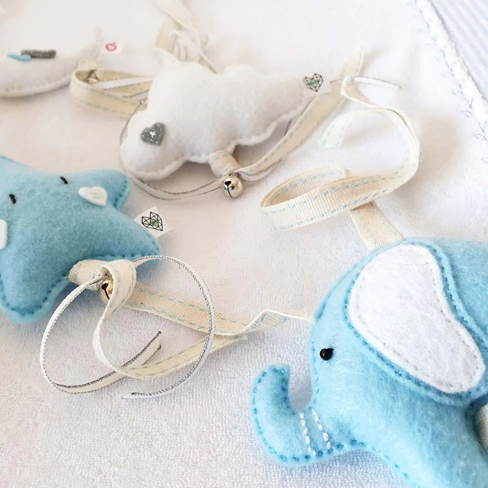 A close-up of soft baby plush toys on a white fabric background, including a light blue elephant, a white cloud ornament, and a pale blue star-shaped toy with cream ribbons.