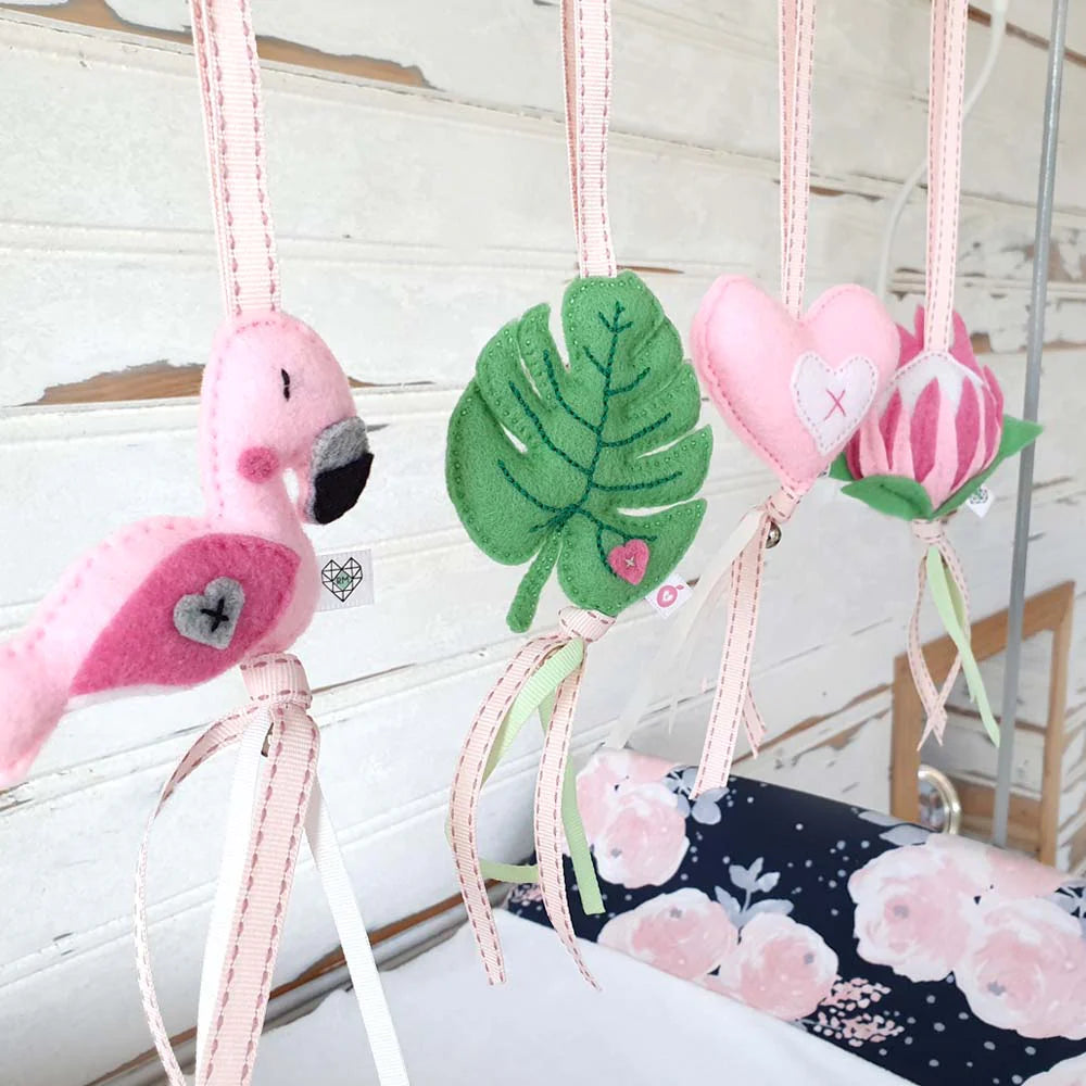 Pink felt baby nursery hanging decorations including a flamingo, green monstera leaf, pink heart, and pink flower, suspended by ribbons against a white wood paneled wall.