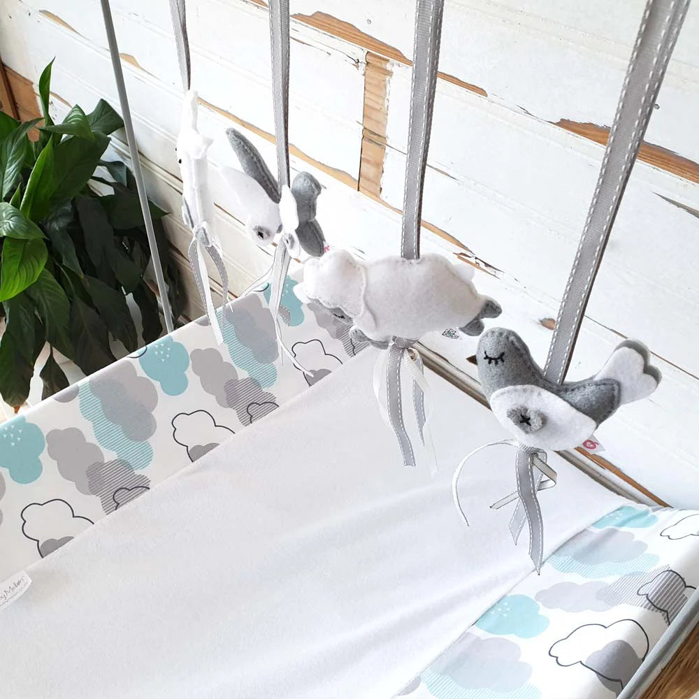 A baby crib bedding set featuring a white fitted sheet and matching cloud-patterned crib bumper and headboard cover in gray and light blue, with a gray and white plush bird mobile hanging above.