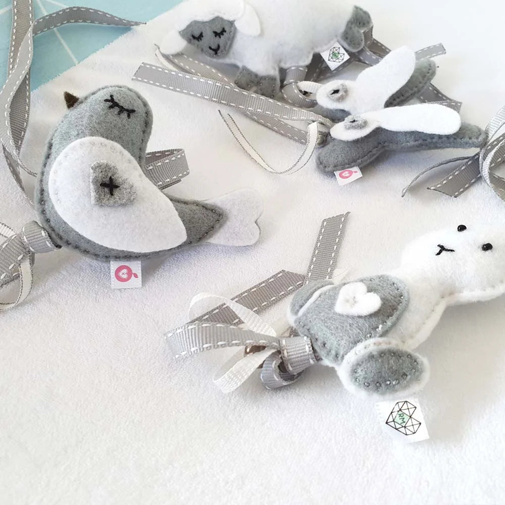 A set of handmade felt animal ornaments, including a seal, a rabbit, and a sheep, crafted from gray and white felt with silver-gray ribbons, displayed on a white textured fabric background.