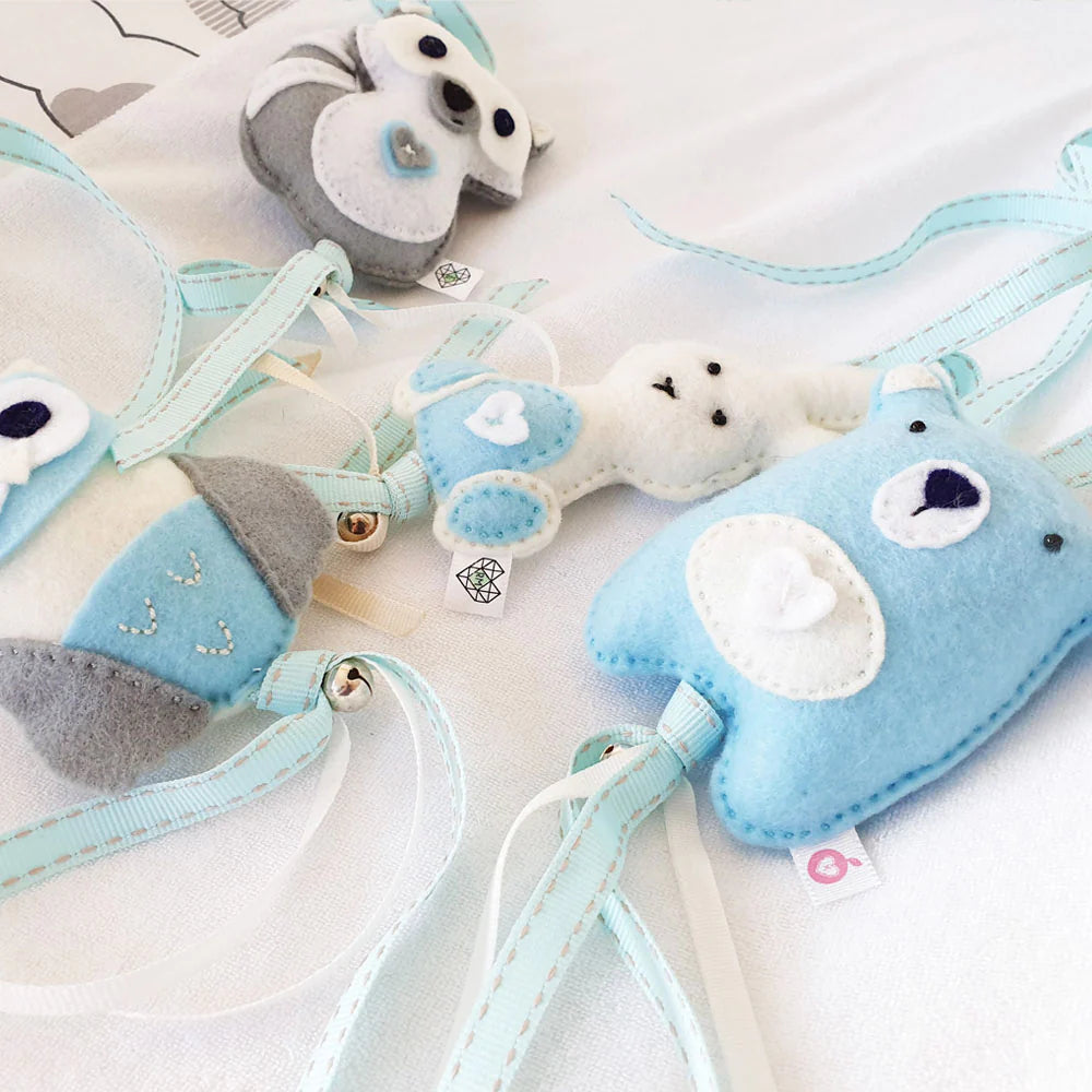 A set of soft felt baby keepsakes or tags shaped like animals, including a blue bear, a white bunny, and a gray-and-white owl, attached to pale blue ribbons on a white fabric background.