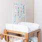 A baby changing table with a white padded top and wooden frame, styled with a hanging mobile made of blue, white, and beige wooden beads and ribbons. A white changing pad is fitted, and baby items are stored in the lower shelf area. | Baby Essentials | All Baby Products | Soft Spot Kids | South Africa