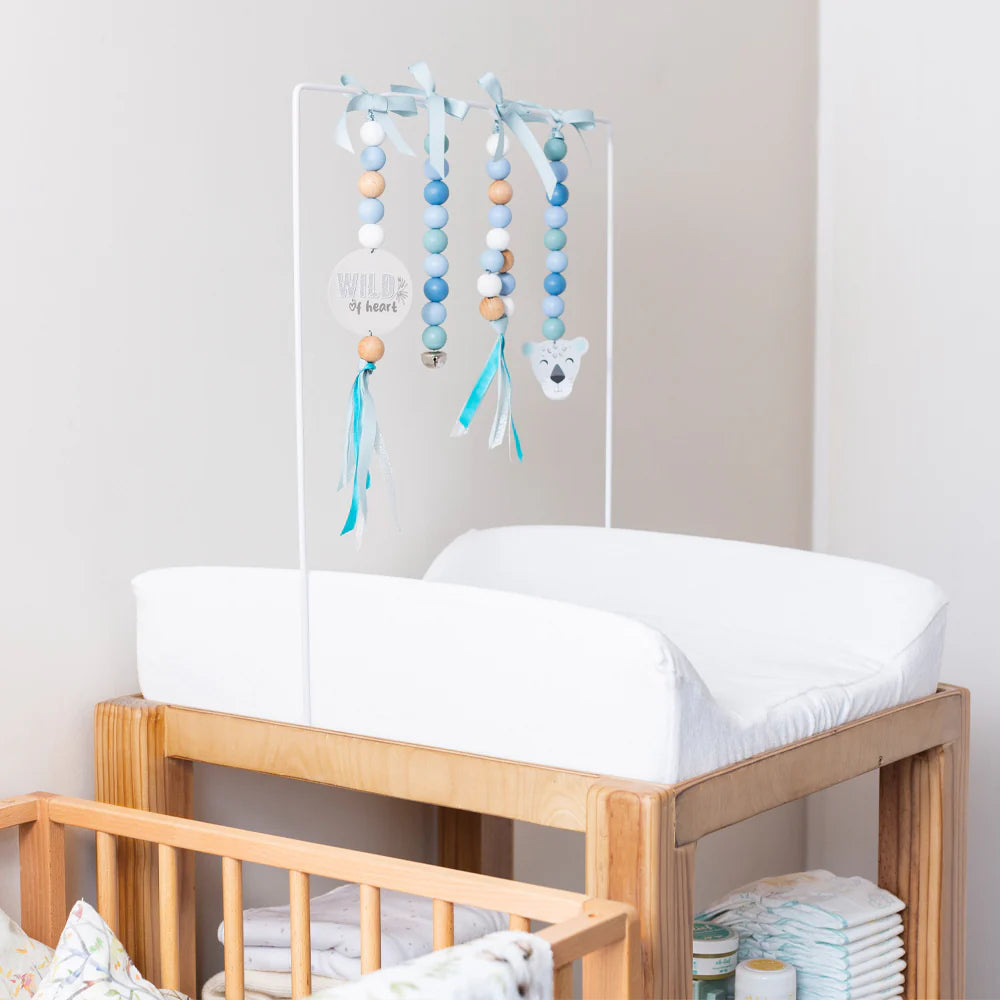 A baby changing table with a white padded top and wooden frame, styled with a hanging mobile made of blue, white, and beige wooden beads and ribbons. A white changing pad is fitted, and baby items are stored in the lower shelf area.