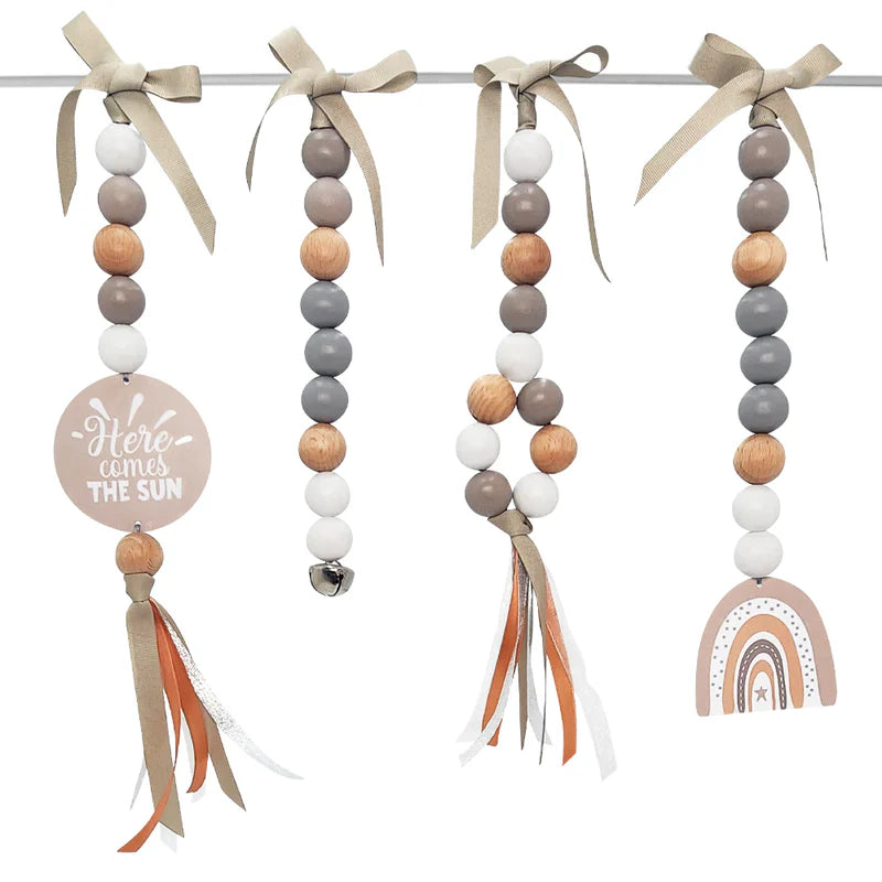 Four boho-style beaded baby wall hangings suspended from a metal rod with beige ribbon bows. The set includes a circular tag reading 'Here comes THE SUN,' a bell bead, a wood-and-white beaded strand with orange ribbon tassels, and a wooden rainbow charm.