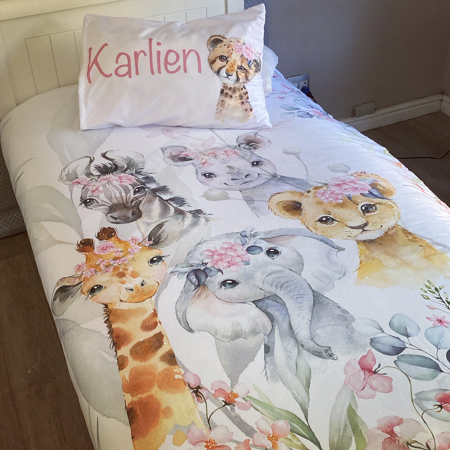 A toddler bedding set with a pink safari theme, featuring cartoonish animal characters such as a giraffe, elephant, and others, along with floral designs, presented on a bed with a child's name 'Karlien' on a pillowcase.