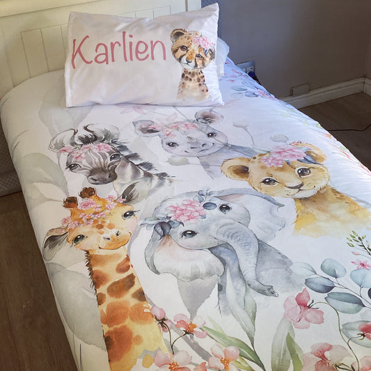 A toddler bedding set with a pink safari theme, featuring cartoonish animal characters such as a giraffe, elephant, and others, along with floral designs, presented on a bed with a child's name 'Karlien' on a pillowcase.