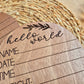 Wooden round card with 'hello world' and fill-in-the-blank sections on a woven mat background. | Baby Essentials | All Baby Products | Soft Spot Kids | South Africa