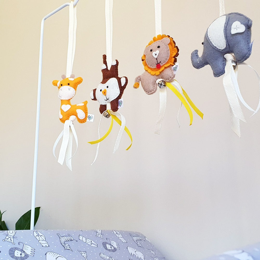 A set of four plush animal baby mobiles hanging from a white metal frame above a bed. The mobiles include a giraffe, a monkey, a lion, and an elephant, each with a different colored ribbon tail.