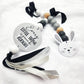 A baby teething ring and lollipop bar set on a white textured fabric background. The set includes a round silver metal rattle with the text 'Every LITTLE THING is gonna be alright', a wooden bead teething ring with black, gray, white, and light wood beads, a white bunny head lollipop, and black and white ribbons. | Baby Essentials | All Baby Products | Soft Spot Kids | South Africa