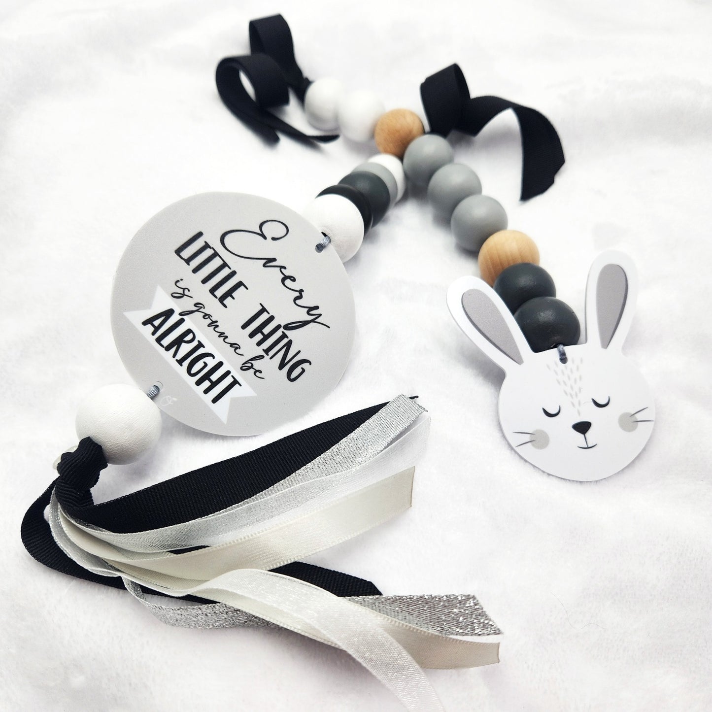 A baby teething ring and lollipop bar set on a white textured fabric background. The set includes a round silver metal rattle with the text 'Every LITTLE THING is gonna be alright', a wooden bead teething ring with black, gray, white, and light wood beads, a white bunny head lollipop, and black and white ribbons.