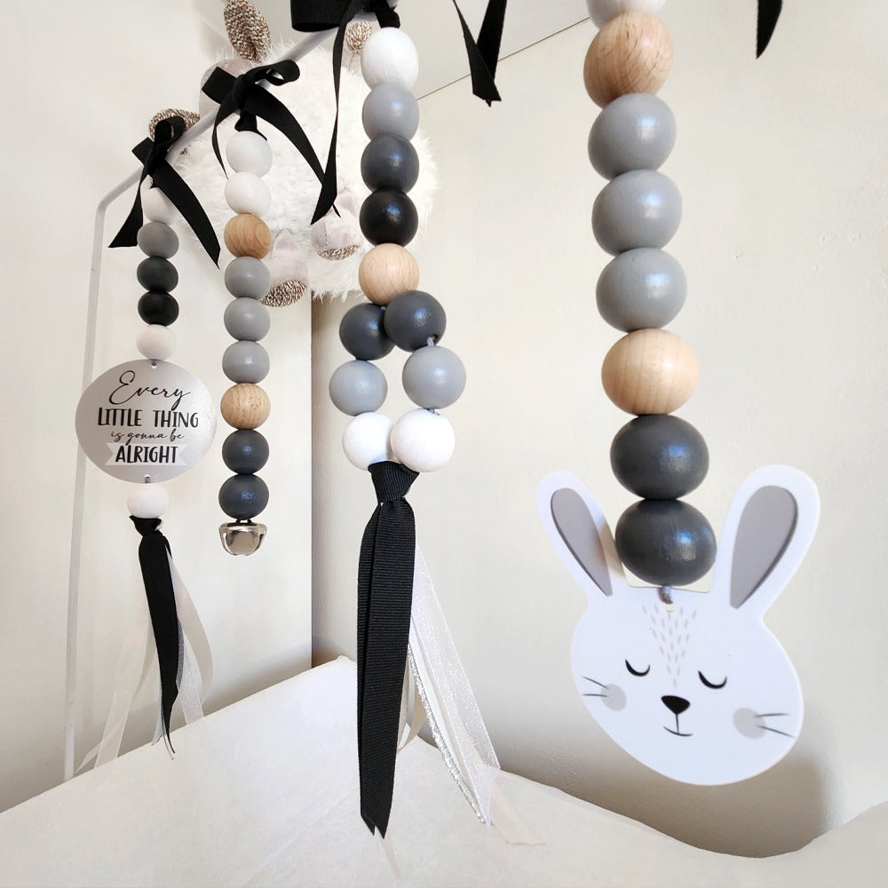 Easter-themed nursery wall hanging with wooden bead garlands, black ribbon bows, a white bunny face cutout, and a round sign that reads 'Every little thing is alright!'.
