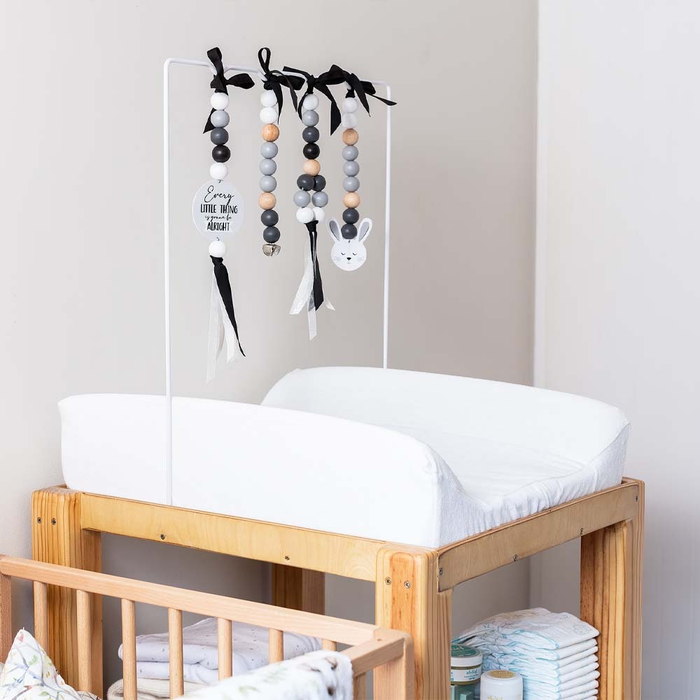 A white changing pad on a wooden changing table in a nursery, with a white mobile hanging above it featuring black ribbons, gray and white beads, and a white bunny decoration.