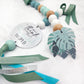 A decorative beaded garland with pastel blue, mint green, white, and natural wood beads, a round silver tag reading 'and I think it wonderful WORLD', a paper monstera leaf charm, and green and turquoise ribbons on a white textured background. | Baby Essentials | All Baby Products | Soft Spot Kids | South Africa