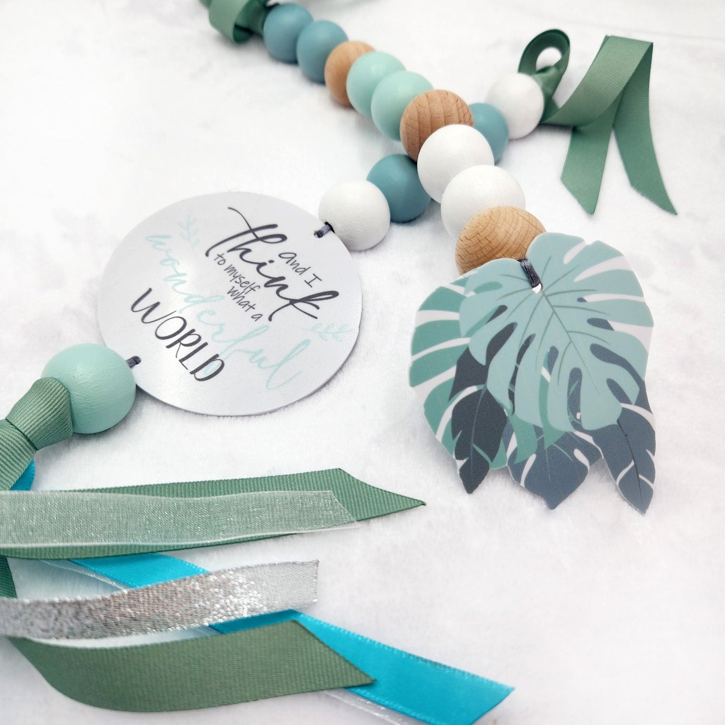 A decorative beaded garland with pastel blue, mint green, white, and natural wood beads, a round silver tag reading 'and I think it wonderful WORLD', a paper monstera leaf charm, and green and turquoise ribbons on a white textured background.