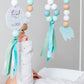A baby's hand holds a mint green and white wooden bead mobile with pastel blue, white, and natural wood beads, ribbons, and hanging tags in a soft gray nursery setting. | Baby Essentials | All Baby Products | Soft Spot Kids | South Africa