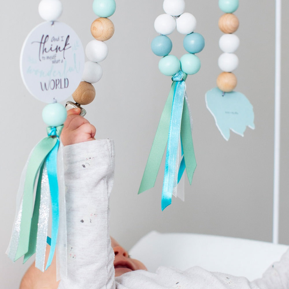 A baby's hand holds a mint green and white wooden bead mobile with pastel blue, white, and natural wood beads, ribbons, and hanging tags in a soft gray nursery setting.