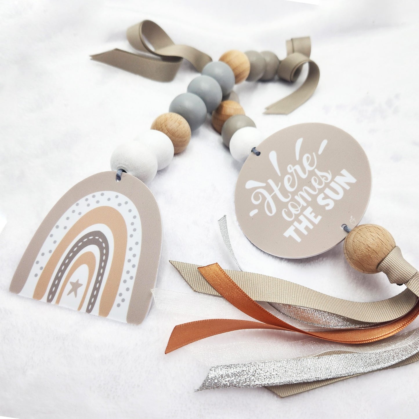 A baby teething toy or pacifier clip made with wooden beads, taupe ribbon, a round tag reading 'Here comes THE SUN,' and a rainbow-shaped tag on a soft white fabric background.
