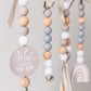A close-up of a hanging beaded baby mobile or garland with pastel wooden beads, white beads, ribbon ties, a round sign reading 'Here comes THE SUN,' and a small rainbow charm visible in the background. | Baby Essentials | All Baby Products | Soft Spot Kids | South Africa