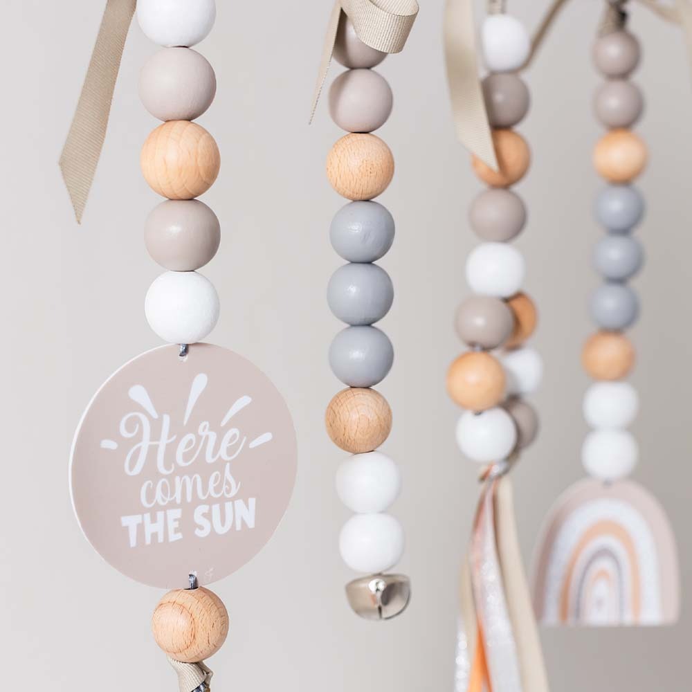 A close-up of a hanging beaded baby mobile or garland with pastel wooden beads, white beads, ribbon ties, a round sign reading 'Here comes THE SUN,' and a small rainbow charm visible in the background.