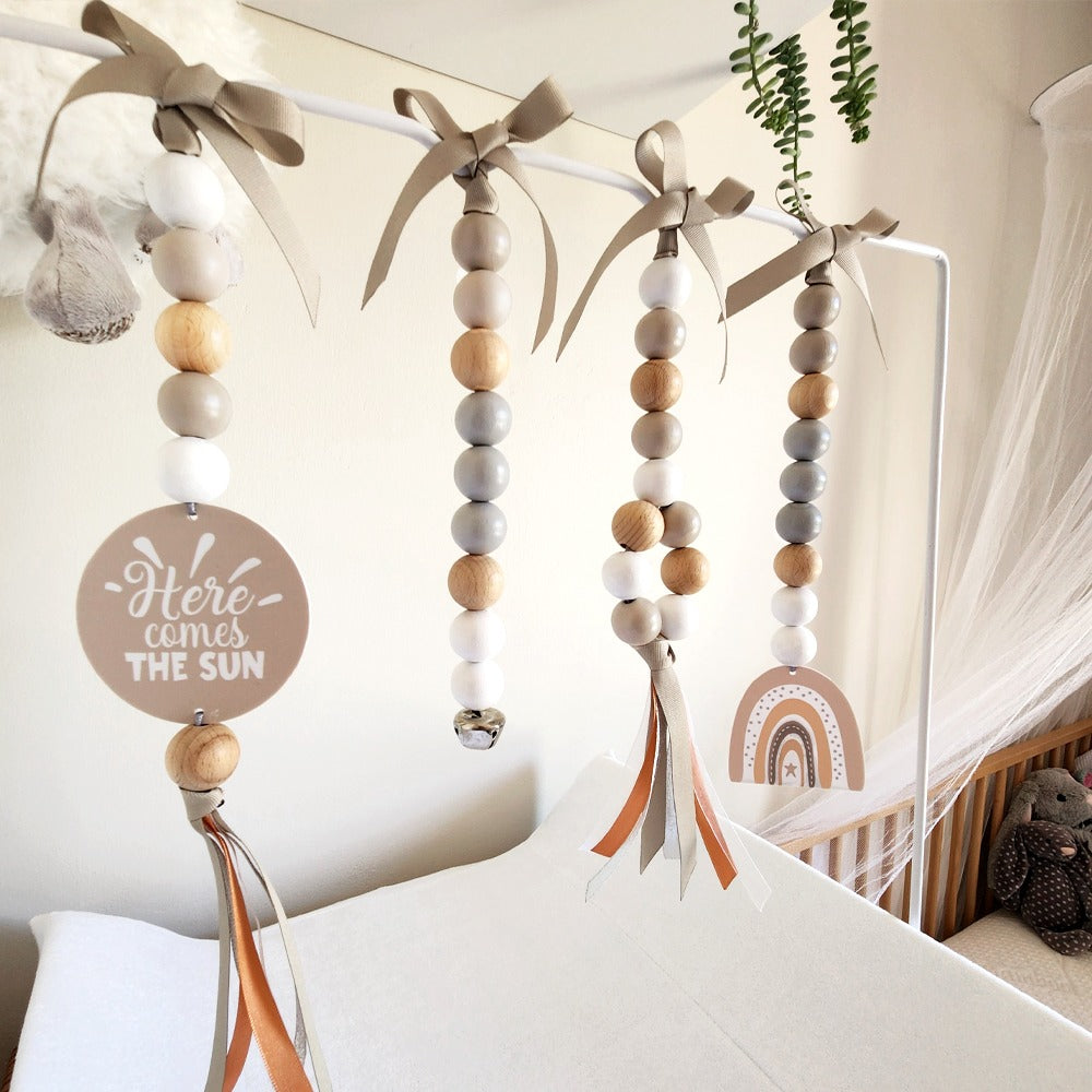 Three wooden bead baby mobiles hang from a white canopy in a nursery. The mobiles feature natural wood, white, and gray beads, tied with taupe ribbons at the top, and decorated with a round wooden sign reading 'Here comes THE SUN' and a small rainbow charm.