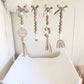 A baby nursery hanging decoration set featuring four vertical beaded garlands with gray, white, and natural wood beads, taupe ribbon bows, tassels, a round wooden sign reading 'Here comes the sun,' and a wooden rainbow ornament, displayed on a white changing table against a light wall. | Baby Essentials | All Baby Products | Soft Spot Kids | South Africa