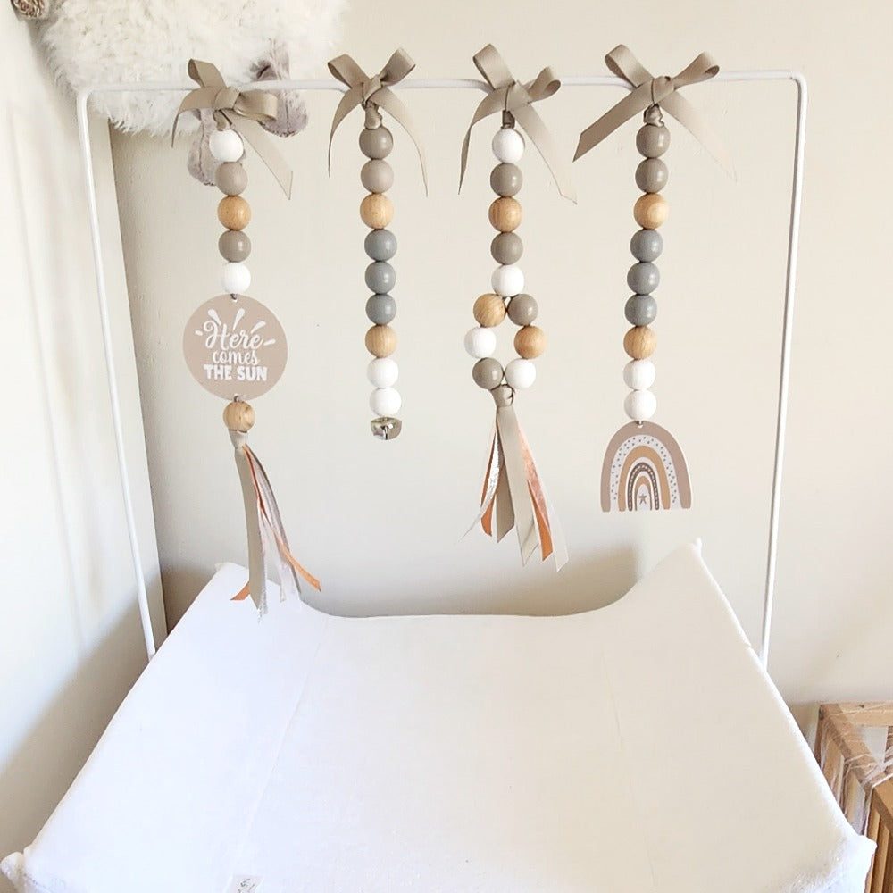 A baby nursery hanging decoration set featuring four vertical beaded garlands with gray, white, and natural wood beads, taupe ribbon bows, tassels, a round wooden sign reading 'Here comes the sun,' and a wooden rainbow ornament, displayed on a white changing table against a light wall.
