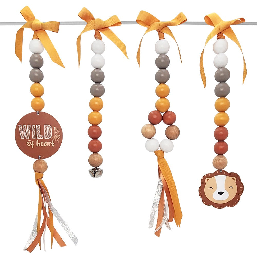Three beaded baby hanging toys on a white background, each with orange and yellow ribbons, wooden beads in orange, brown, white, and gray, and decorative elements including a round 'WILD of heart' sign and a lion face.