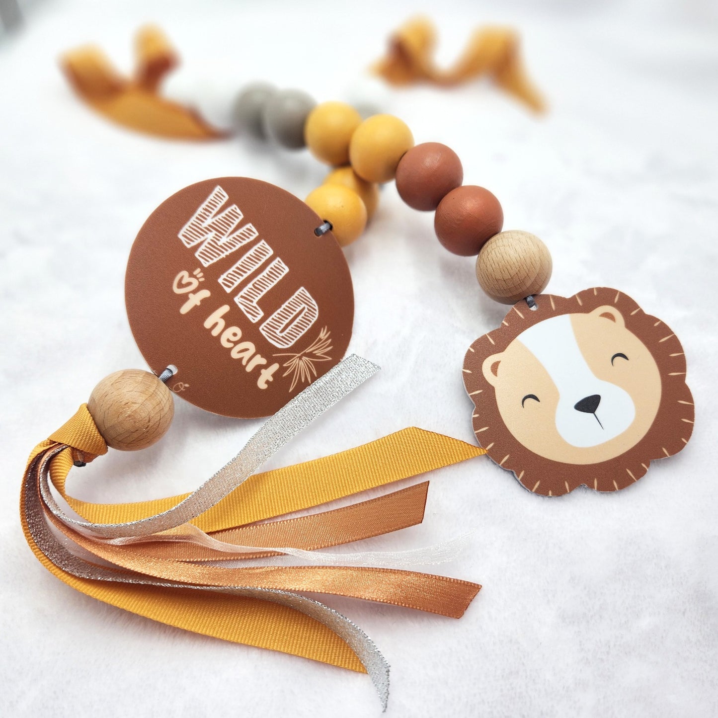 A decorative baby keepsake or gift tag featuring a lion face charm, wooden beads, yellow and brown ribbons, and a round brown tag that reads 'Wild of heart' on a soft white textured background.