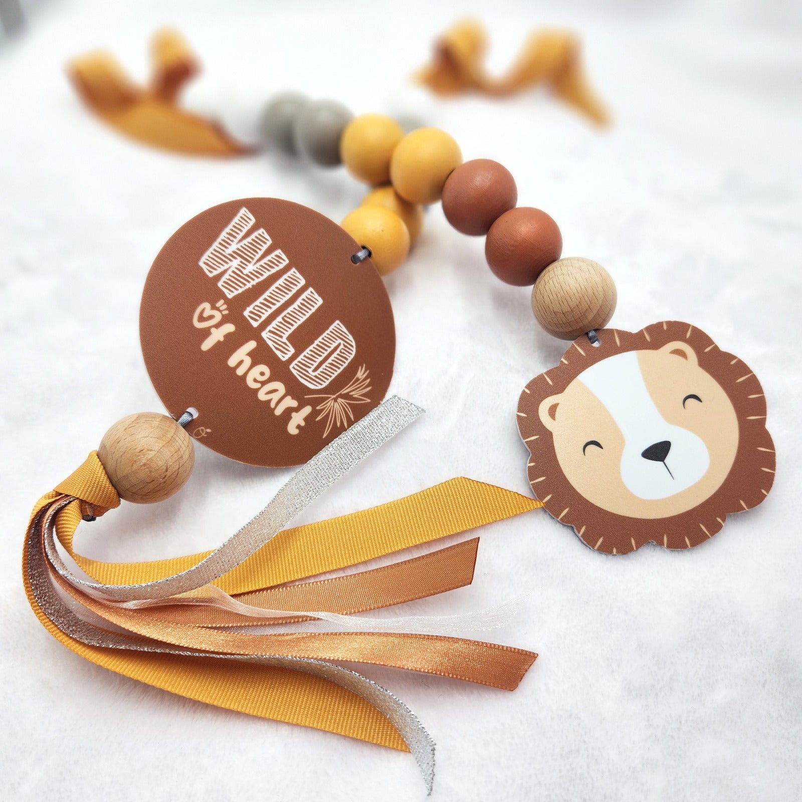 A decorative baby keepsake or gift tag featuring a lion face charm, wooden beads, yellow and brown ribbons, and a round brown tag that reads 'Wild of heart' on a soft white textured background.