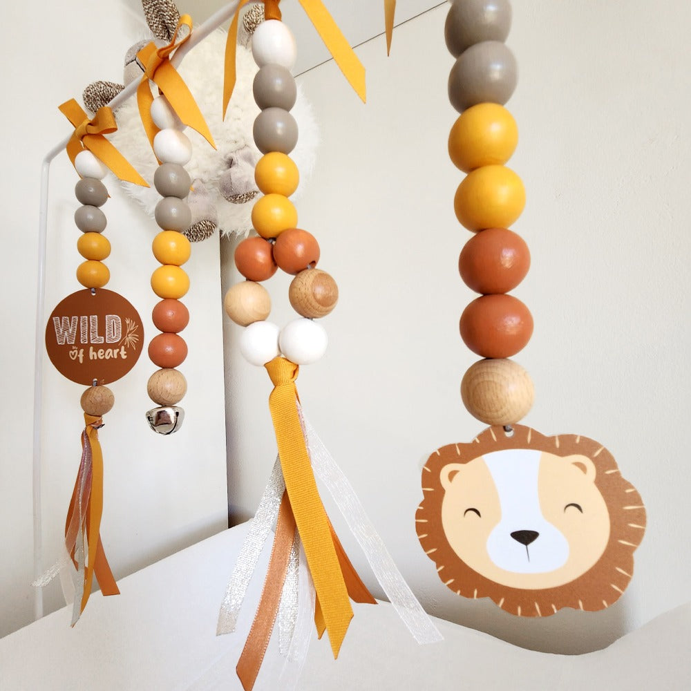 Wooden hanging baby wall decor with colorful beads, yellow and orange ribbons, a round 'WILD of heart' sign, and a lion face cutout on a white wall.