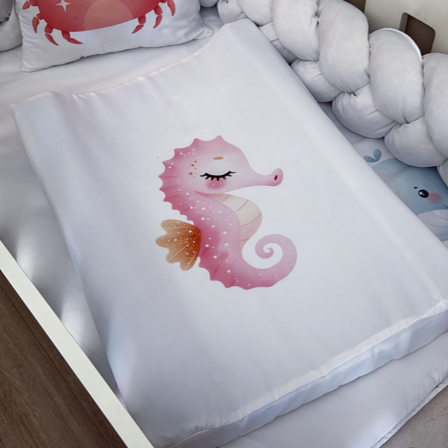 White pillow with a pink seahorse design on a bed with white pillows and a wooden sideboard.