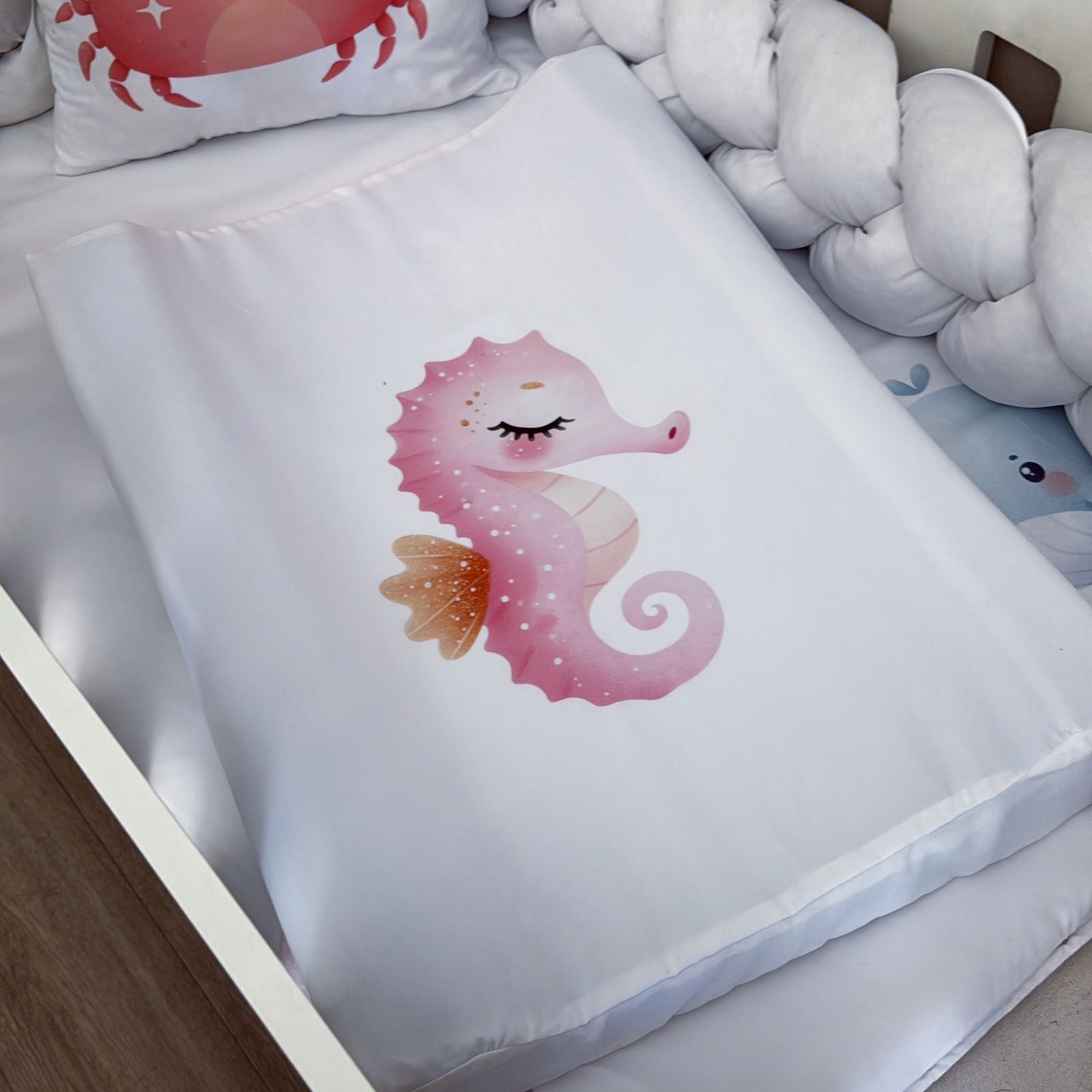 White pillow with a pink seahorse design on a bed with white pillows and a wooden sideboard.