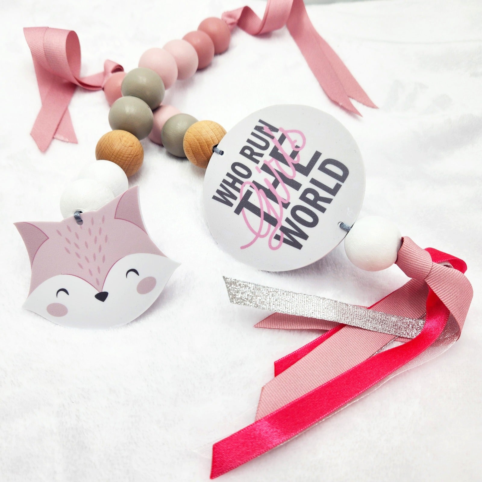 A baby keepsake beaded necklace with pink and peach wooden beads, white beads, a pink fox head charm, a round white tag reading 'WHO RUN THE WORLD', and pink and red ribbons on a white fabric background.