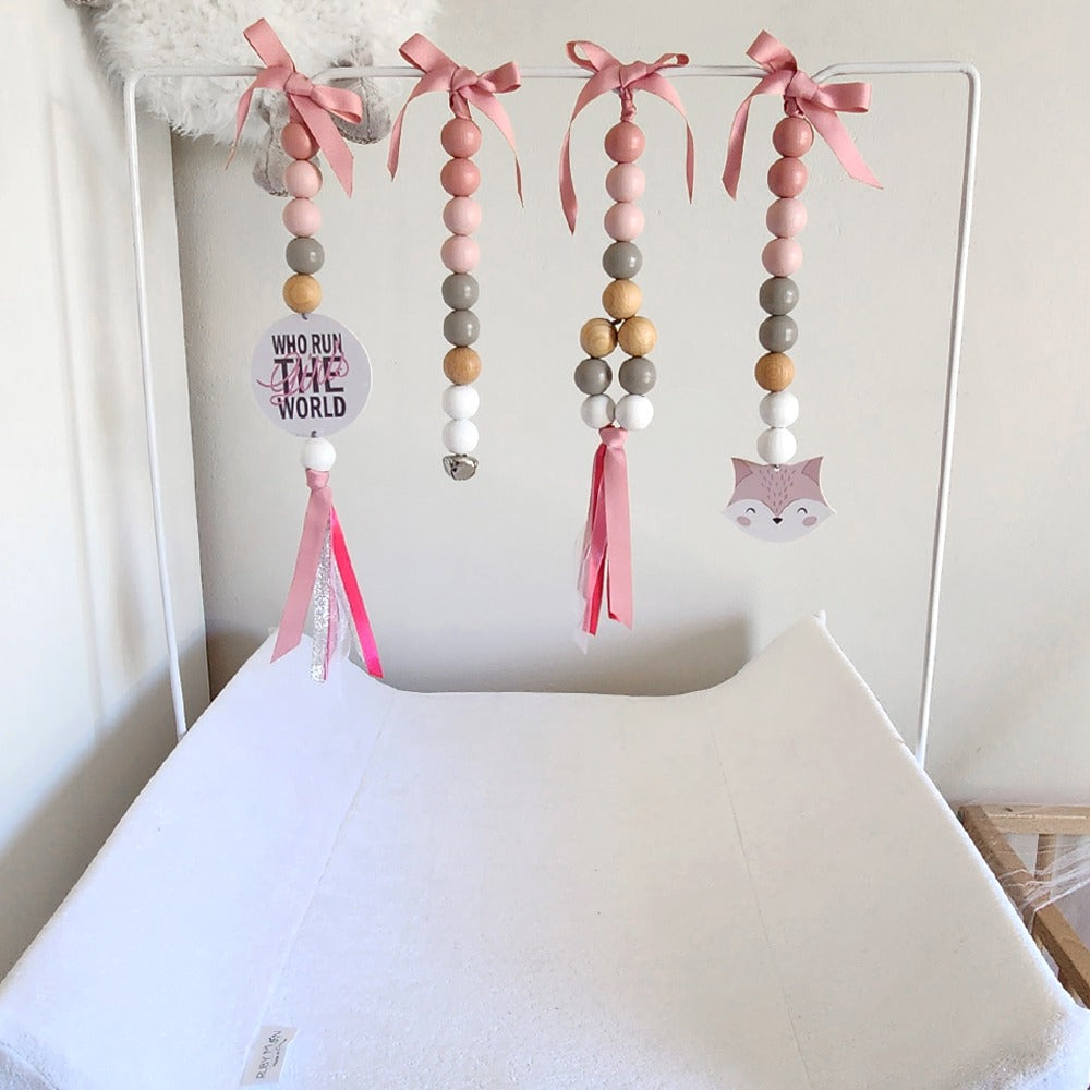 A pink and neutral wooden bead baby mobile hanging from a white metal frame above a white changing pad. The mobile features four vertical strands of beads in shades of pink, beige, gray, and white, accented with pink ribbons, a small 'WHO WANTS THE WORLD' sign, and a pink fox head charm.