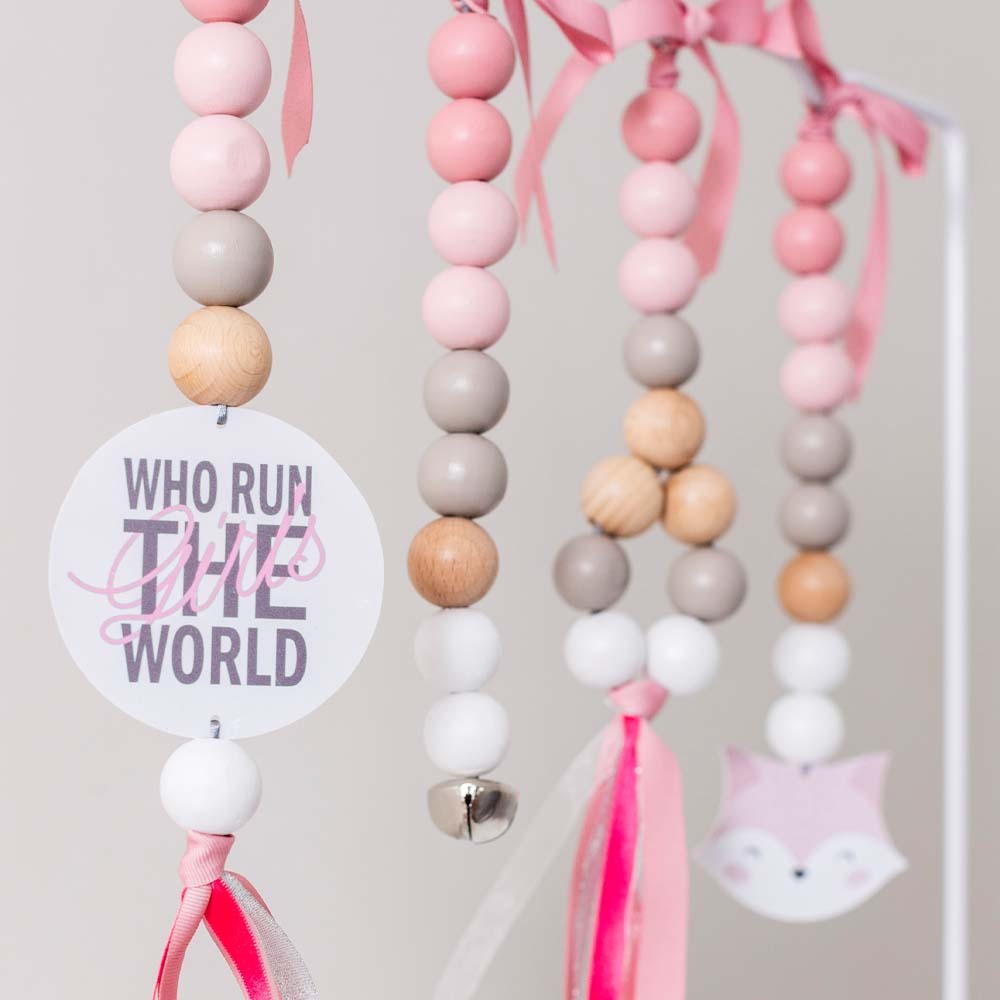 A close-up of a hanging beaded decoration with pastel pink, white, and light wood beads, pink ribbons, a round white tag reading 'WHO RUN THE WORLD,' and a small pink fox face charm.