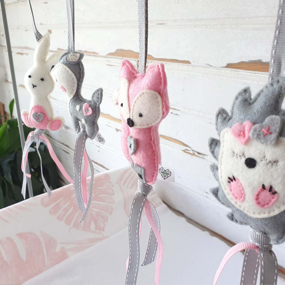 A set of handmade felt animal hanging ornaments, including a pink fox, a white bunny, and a gray hedgehog, decorated with pink and gray ribbons against a rustic white wooden background.