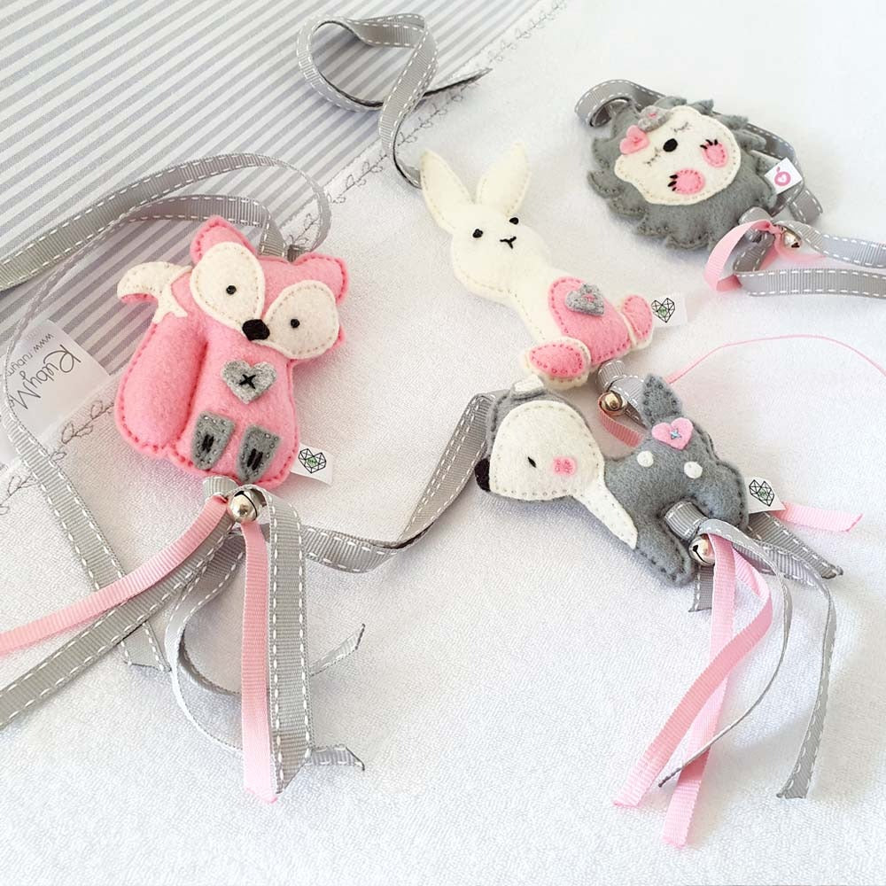 A set of four handmade felt animal charms with silver and pink ribbons, including a pink fox, white bunny, gray lion, and gray elephant, arranged on a white textured background.