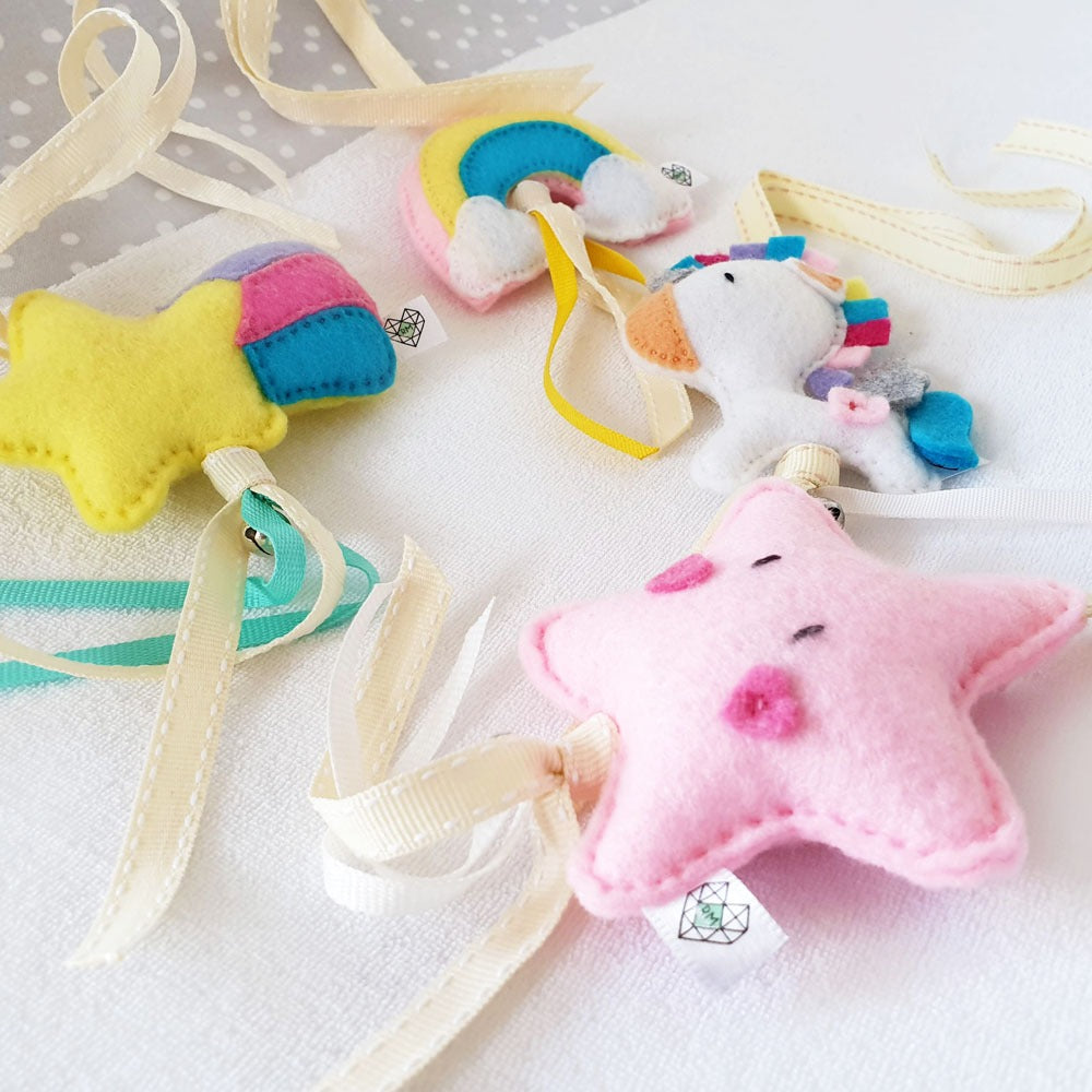 A set of handmade felt baby accessories including a pink star with embroidered eyes and mouth, a yellow star with pink and blue accents, a rainbow, and a unicorn, all attached to cream-colored ribbons on a white fabric background.