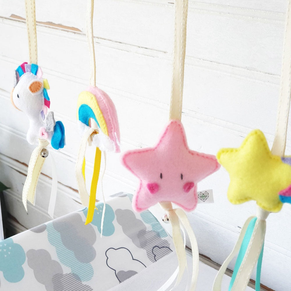 A pastel baby mobile with soft felt toys hanging in front of a white paneled wall. The mobile includes a white unicorn with rainbow mane, a pink-and-yellow rainbow, a pink star face, and a yellow star face, suspended by cream ribbons over a cloud-patterned fabric.